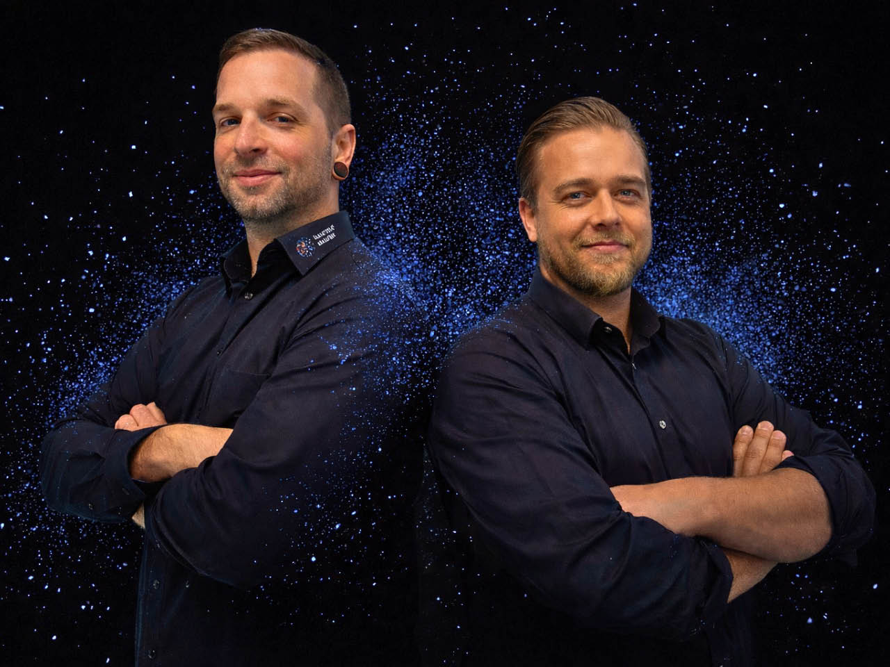 Zwei Männer in schwarzen Hemden stehen mit verschränkten Armen vor dunklem Hintergrund mit blauen Partikeleffekten und blicken freundlich in die Kamera